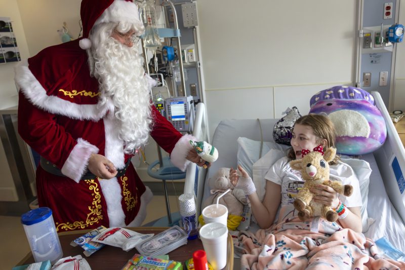 A young girl in a hospital bed, in a room filled with medical equipment and other supplies, smiles as she reached out to accept a gift of a small stuffed animal from Santa Claus.