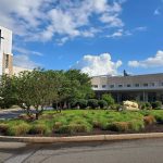 Front entrance of Penn State Health St. Joseph Medical Center with a landscaped circular driveway, a grassy area featuring small bushes, trees, and a statue of the nittany lion. The building has a tall structure with a cross, large glass windows, and a blue sky with scattered white clouds in the background.