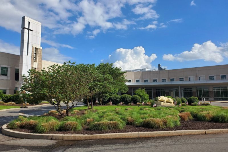 Front entrance of Penn State Health St. Joseph Medical Center with a landscaped circular driveway, a grassy area featuring small bushes, trees, and a statue of the nittany lion. The building has a tall structure with a cross, large glass windows, and a blue sky with scattered white clouds in the background.