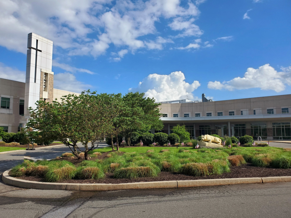 Front entrance of Penn State Health St. Joseph Medical Center with a landscaped circular driveway, a grassy area featuring small bushes, trees, and a statue of the nittany lion. The building has a tall structure with a cross, large glass windows, and a blue sky with scattered white clouds in the background.