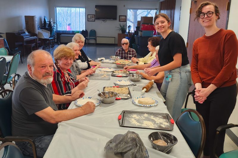 A group of older adults and medical students stands and sits at a table during a cooking class.