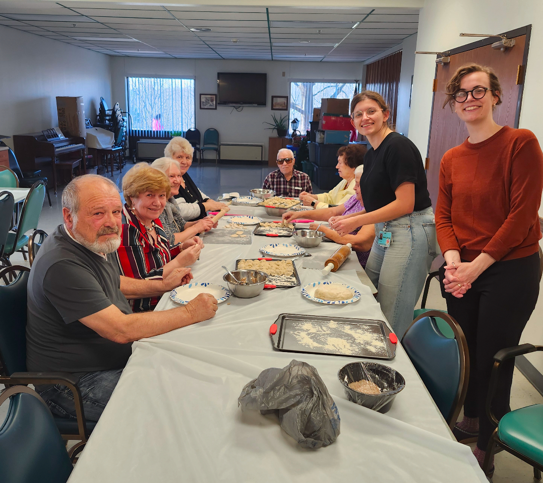 A group of older adults and medical students stands and sits at a table during a cooking class.