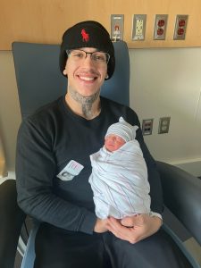 A man smiles as he sits in a chair in a hospital room, holding a swaddled infant.