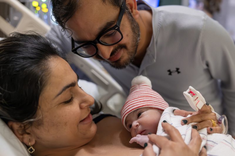 A baby lays on her mother's chest while dad crouches by their hospital bed.