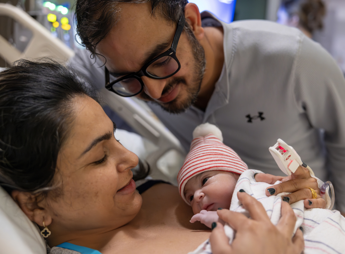 A baby lays on her mother's chest while dad crouches by their hospital bed.