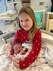 A young girl smiles as she sits on a hospital bed holding her baby sister.