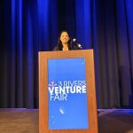 A woman stands on a stage, at a podium, to pitch a business idea.