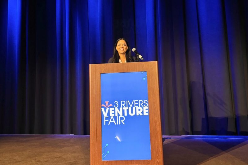 A woman stands on a stage, at a podium, to pitch a business idea.