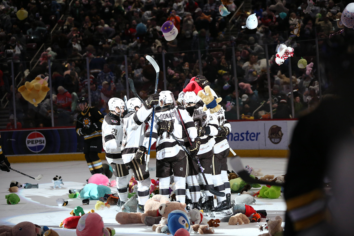 A group of professional hockey players gather facing each other on a hockey rink. Stuffed animals are on the ice around them and flying through the air from the spectators who are throwing them.