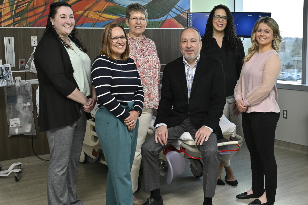 Six people in business casual attire pose for a photo in a hospital room. A hospital bed, computer and other medical equipment are in the background.