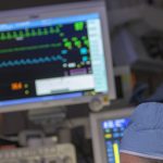 The top of a medical provider’s head, wearing a surgical cap, is in the foreground. A monitor with a patient’s vital signs is in the background, slightly out of focus.