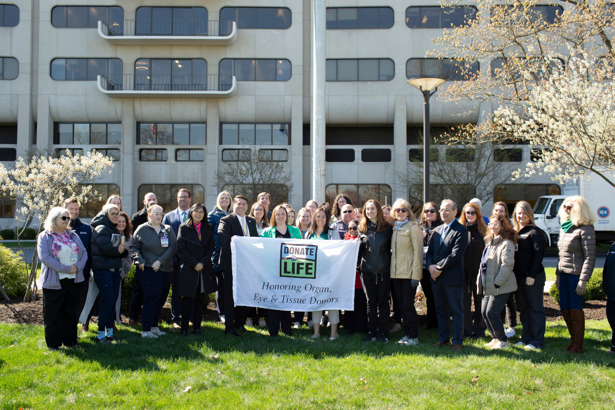 A group of people pose with a “Donate Life” flag. A hospital building with windows is in the background.