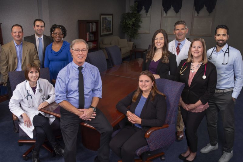 A group of male and female physicians and physician assistants, wearing professional attire, stand and sit in a group photo.