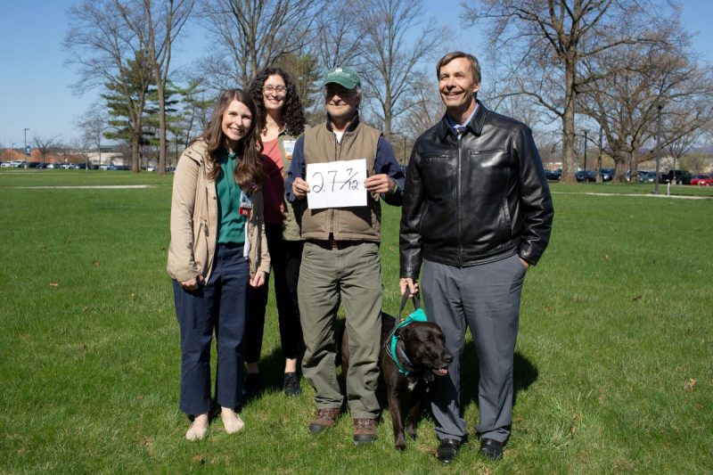 Two women and two men smile as they pose for a photo outdoors on the Milton S. Hershey Medical Center campus. The man in the middle is holding a sign that reads 27-7/12. The man on the right is holding a service dog’s collar.