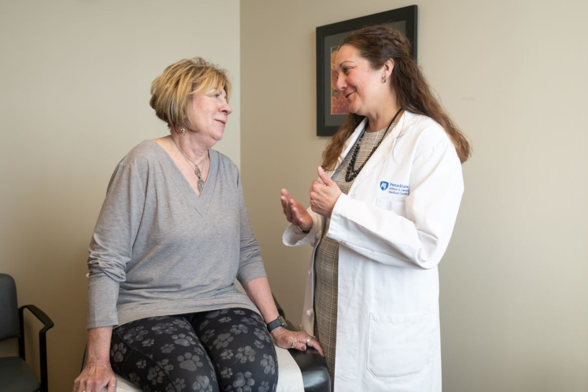 A woman sits on an exam table talking to Dr. Jaime Long, right, of Penn State Health. Dr. Long is gesturing with her hands and wearing a white coat with the Penn State Health Milton S. Hershey Medical Center logo on it. Both are smiling.