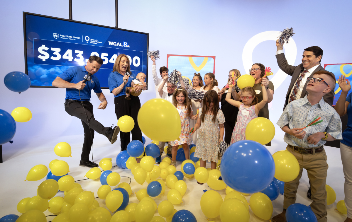 A group of excited children and adults celebrate on stage as blue and yellow balloons fall around them. Two WGAL 8 news anchors, one holding a baby, cheer in front of a large screen displaying a fundraising total of $343,054.00.