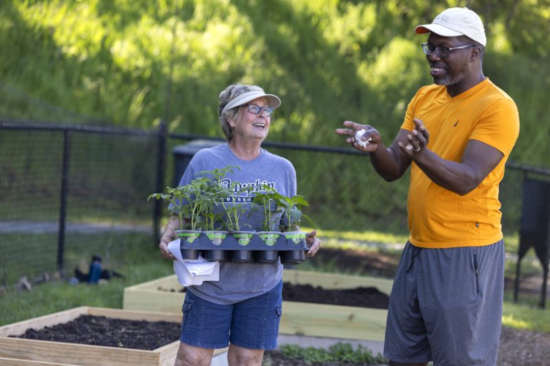 A man and a woman stand side-by-side talking in a community garden. The woman is holding a container with several potted plants. Both individuals are smiling.