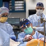 Three people in full surgical attire perform a complex procedure in an operating room. The central surgeon, wearing magnifying surgical loupes and a headlamp, focuses intently on the patient, whose body is draped in sterile coverings.