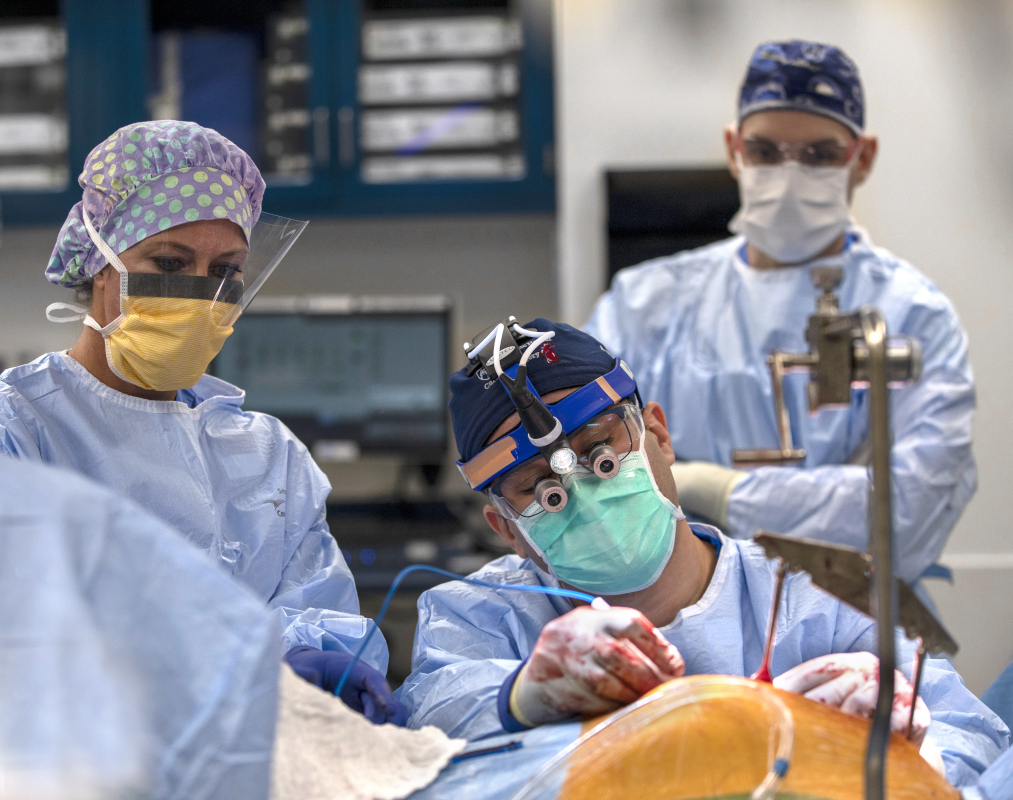 Three people in full surgical attire perform a complex procedure in an operating room. The central surgeon, wearing magnifying surgical loupes and a headlamp, focuses intently on the patient, whose body is draped in sterile coverings.