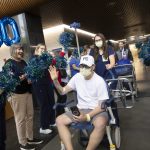 A man wearing a white t-shirt, white baseball cap, and face mask sits in a wheelchair and waves while holding a phone in his lap. He is surrounded by smiling hospital staff wearing scrubs and holding blue and green pom-poms.