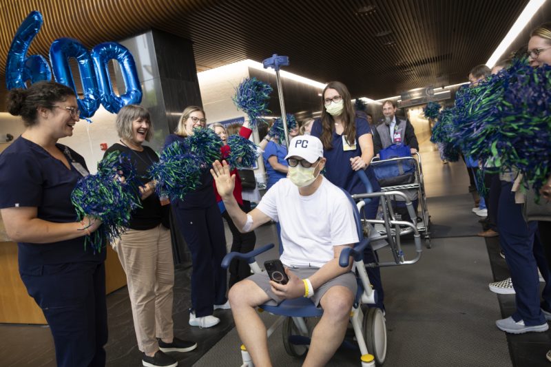 A man wearing a white t-shirt, white baseball cap, and face mask sits in a wheelchair and waves while holding a phone in his lap. He is surrounded by smiling hospital staff wearing scrubs and holding blue and green pom-poms.