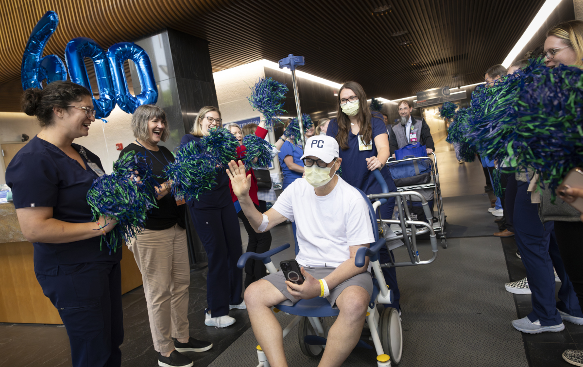 A man wearing a white t-shirt, a white baseball cap and a facial mask is in a wheelchair and waves while holding a phone on his knees. It is surrounded by a smiling hospital staff carrying scrubs and holding blue and green pompoms.