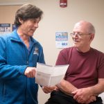 A doctor smiles at a male patient as he points to a paper he is holding. The doctor is wearing a sweat jacket with a Penn State logo on it. The male is wearing a T-shirt and glasses.
