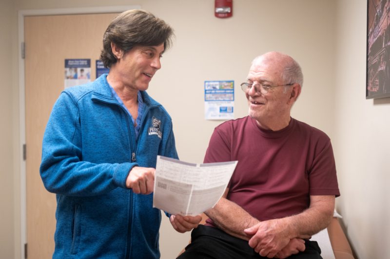 A doctor smiles at a male patient as he points to a paper he is holding. The doctor is wearing a sweat jacket with a Penn State logo on it. The male is wearing a T-shirt and glasses.