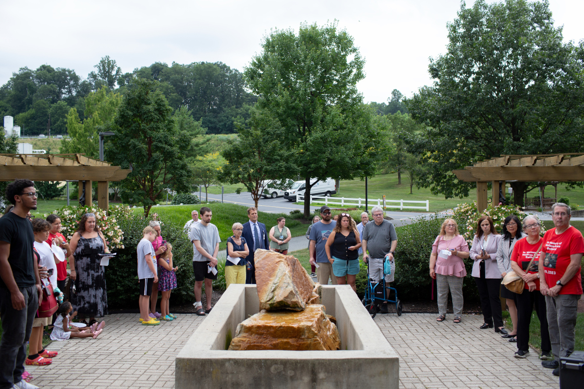 Approximately 20 people stand on a brick patio, surrounding a stone memorial, at a ceremony.
