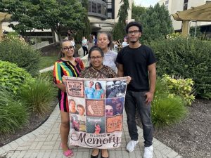 Four people pose for a photo. The woman in front displays a quilt with photos and the words “In Loving Memory.” Bushes are in the background, as is a large building.
