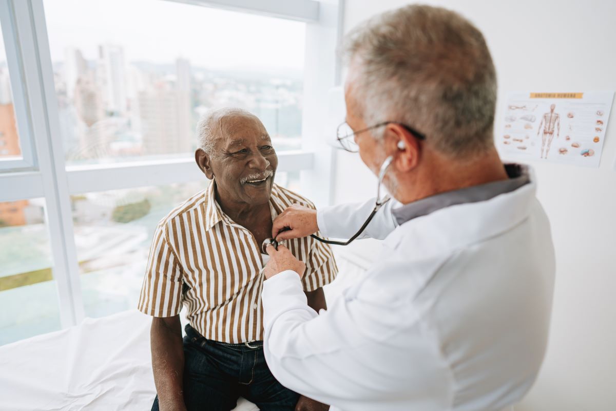 A gentleman is sitting on an exam table while a doctor checks his heart with a stethoscope.