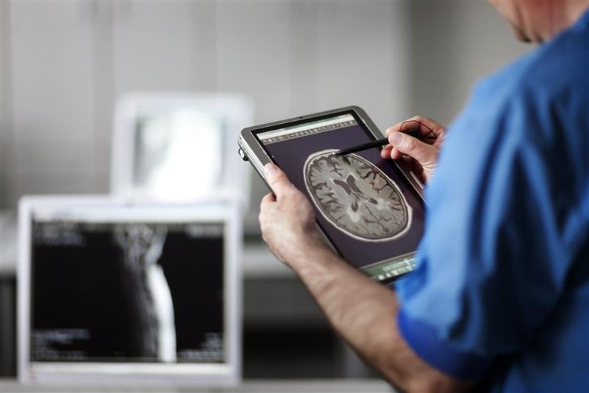 A healthcare professional in blue scrubs examines a brain scan on a digital tablet using a stylus.