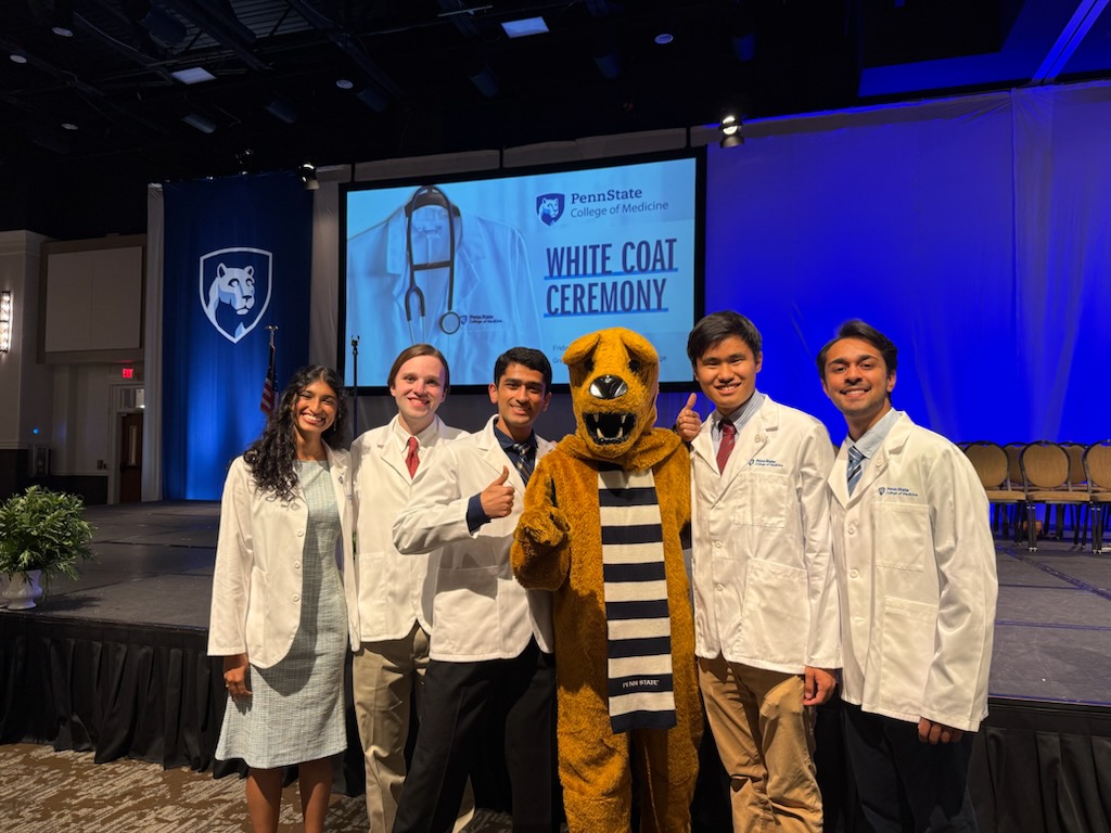 A group of five college students wearing white medical coats stand together in front of a stage.