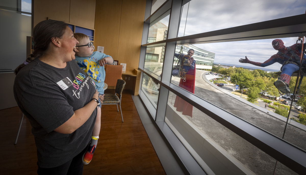 A woman holds a young child in a hospital room as they smile and look out the window. Outside, two window washers dressed as superheroes—Superman and Spider-Man—wave and gesture toward them.