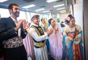 A group of medical students dressed in an array of cultural attire stands together clapping their hands.