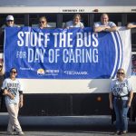 Five people looking out the windows of a bus hold a banner that reads: “Stuff the Bus for Day of Caring.” Three women stand on either side of the banner.