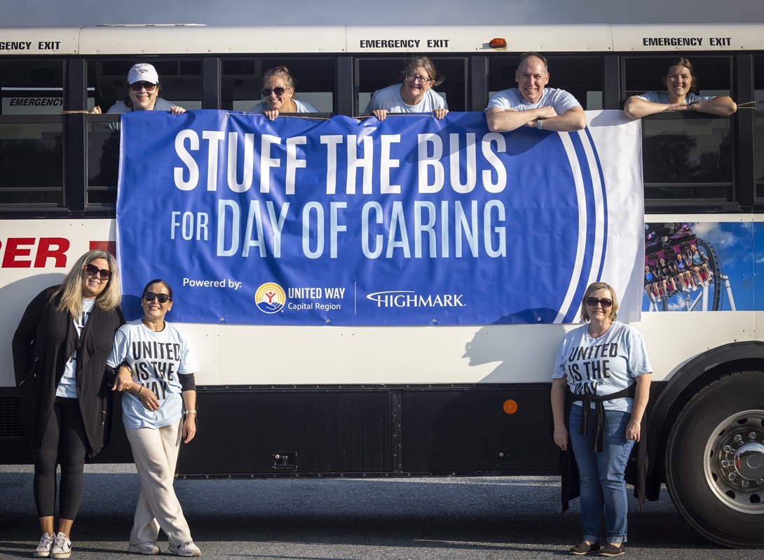 Five people looking out the windows of a bus hold a banner that reads: “Stuff the Bus for Day of Caring.” Three women stand on either side of the banner.