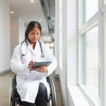 A woman in a wheelchair by a window, wearing a doctor's coat and a stethoscope looks at a digital tablet.
