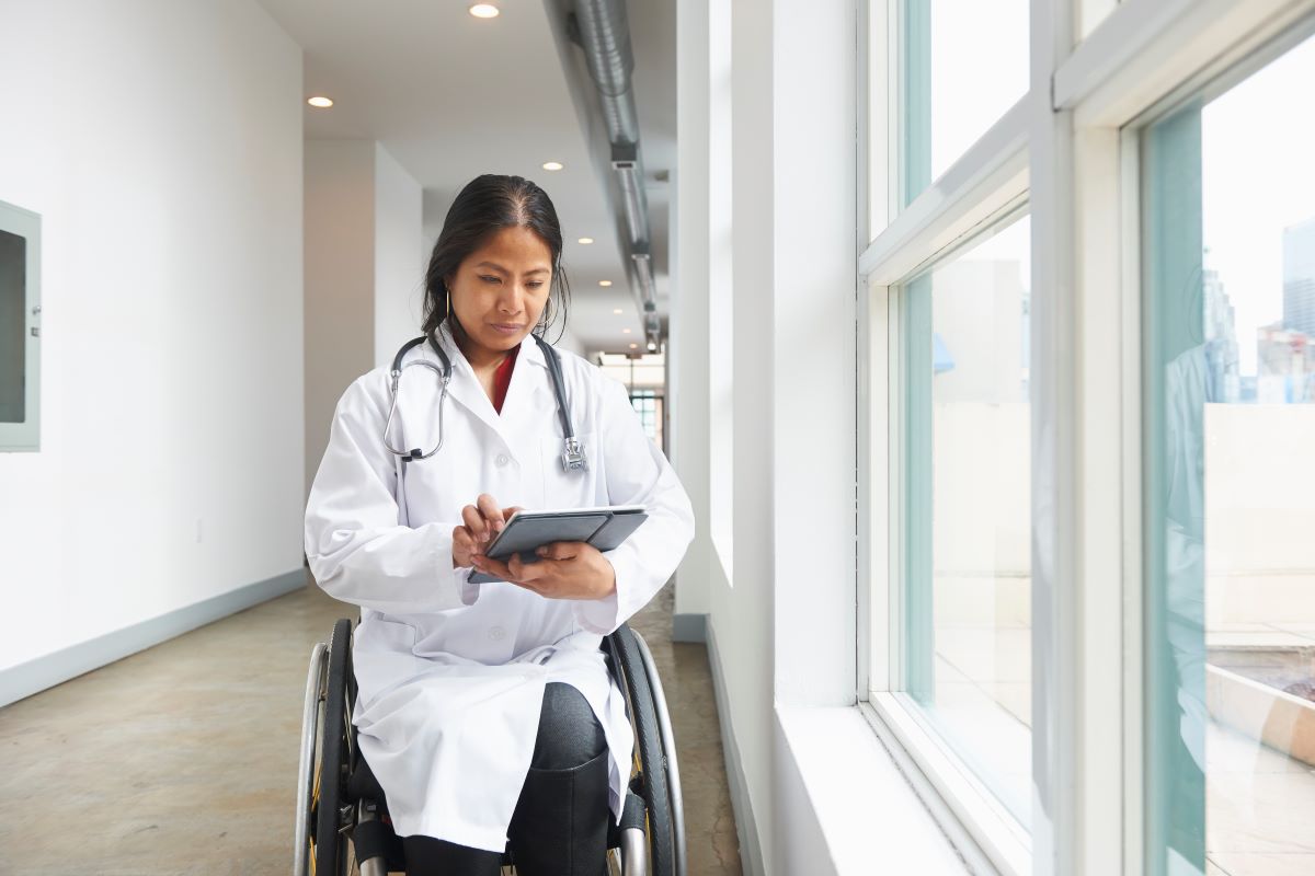 A woman in a wheelchair by a window, wearing a doctor's coat and a stethoscope looks at a digital tablet.
