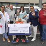 Five people hold up one of their hands toward the camera as they pose for a photo with a check from Hyundai Hope on Wheels for $400,000. A car is in the background, and behind that is Penn State Health Children’s Hospital.