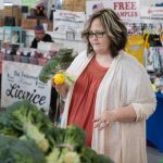 A woman holds a head of broccoli and an orange while looking at other produce at a farmer’s market. She is wearing a sweater and has glasses.