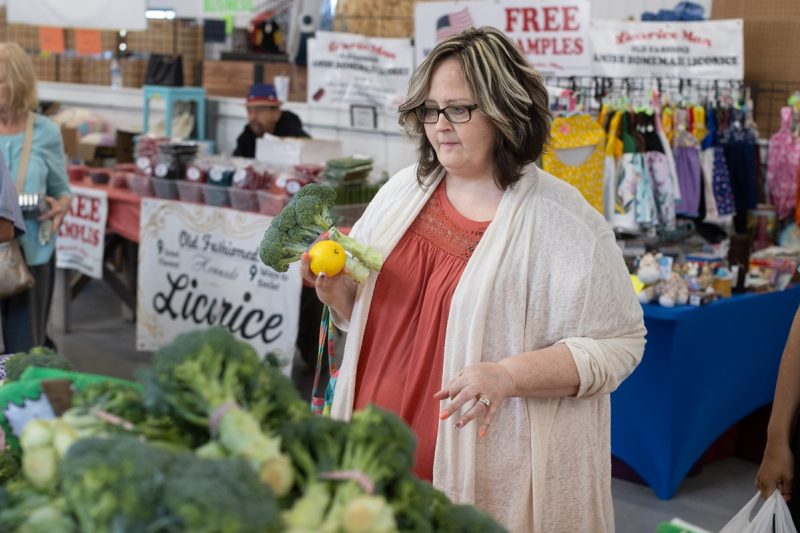A woman holds a head of broccoli and an orange while looking at other produce at a farmer’s market. She is wearing a sweater and has glasses.
