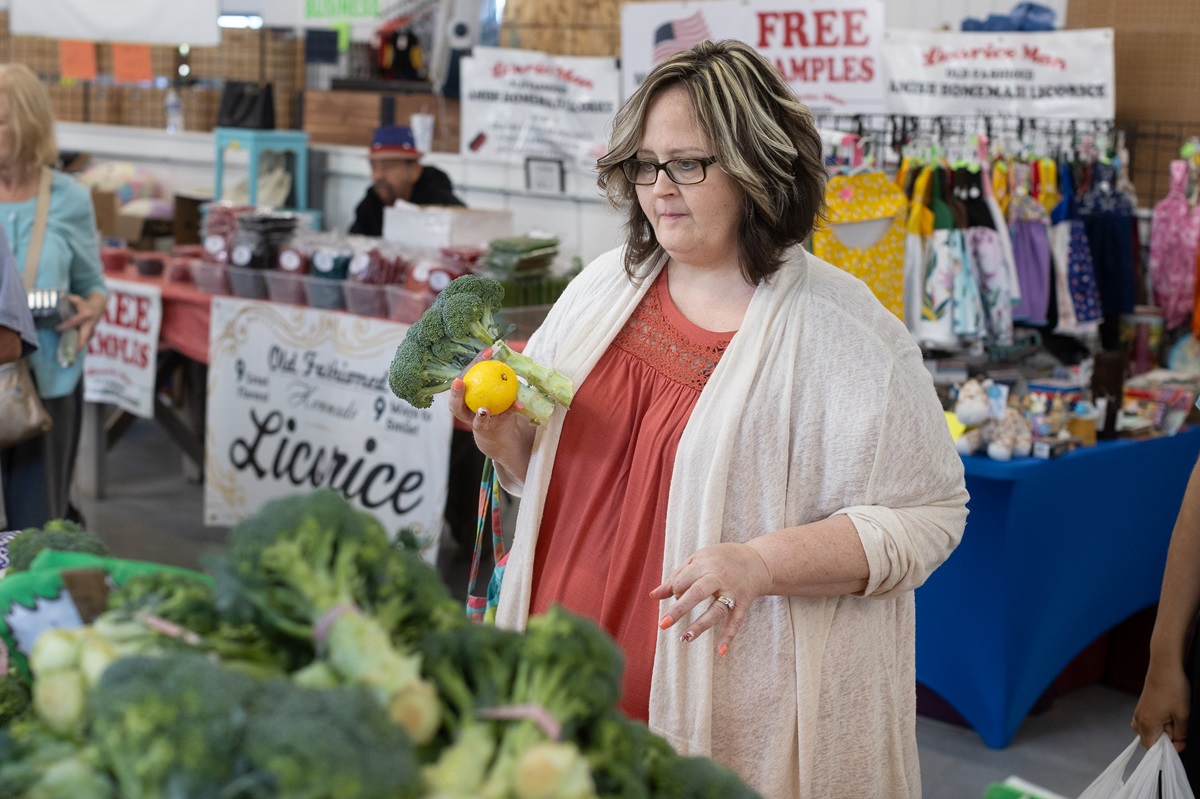 A woman holds a head of broccoli and an orange while looking at other produce at a farmer’s market. She is wearing a sweater and has glasses.
