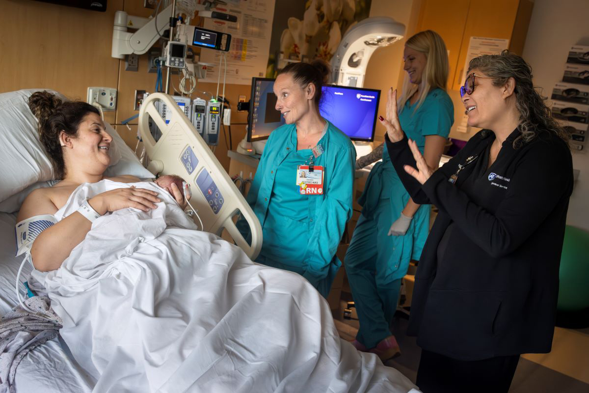A woman lies in a hospital bed, holding a baby. A nurse speaks to the woman while the woman looks at another woman who is interpreting for her in sign language.