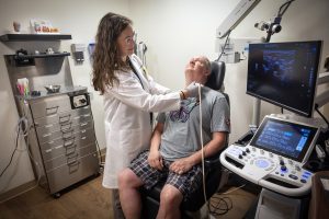 Dr. Emily Funk of Penn State Health Otolaryngology – Head and Neck Surgery uses a neck ultrasound on a male patient as he looks up. The image of his neck appears in a monitor to their right. She is wearing a white lab coat and glasses. He is wearing a T-shirt and shorts.