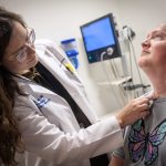 Dr. Emily Funk of Penn State Health Otolaryngology – Head and Neck Surgery points to a scar on the neck of a male patient in an exam room. She is wearing a white lab coat and glasses. He is wearing a T-shirt. Behind them is a monitor.