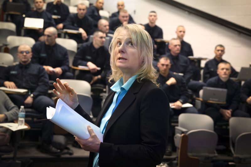Terry Brosche of Penn State Health stands in front of a group of Pennsylvania State Police cadets who are sitting in an auditorium. She is gesturing, looking up and holding papers. She is wearing a suit jacket and blouse.