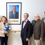 Richard Legro, MD; Cecile Mortel; Joel Sorosky, MD; Raymond Hohl, MD, PhD; and Mark Shahin, MD, stand together smiling beside a portrait of longtime College of Medicine faculty member and humanitarian Rodrigue Mortel, MD, at Penn State Cancer Institute.