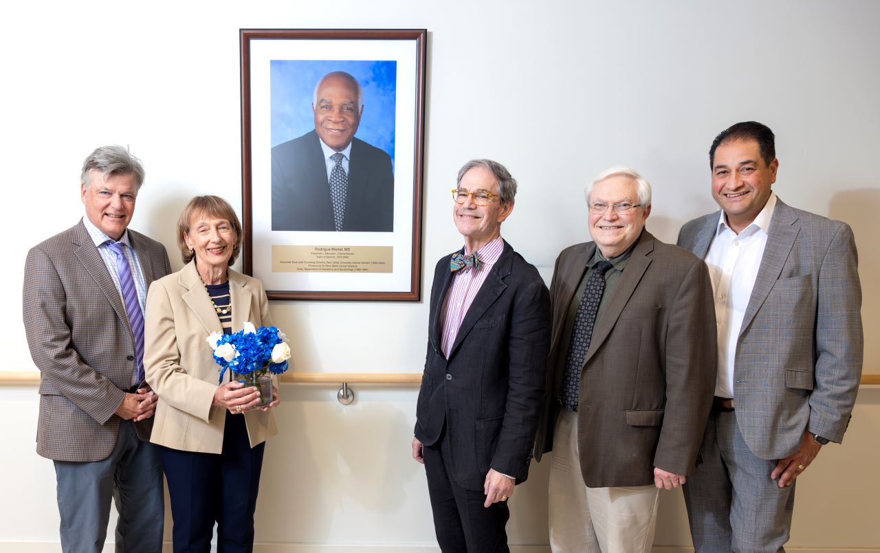 Richard Legro, MD; Cecile Mortel; Joel Sorosky, MD; Raymond Hohl, MD, PhD; and Mark Shahin, MD, stand together smiling beside a portrait of longtime College of Medicine faculty member and humanitarian Rodrigue Mortel, MD, at Penn State Cancer Institute.