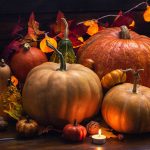 Pumpkins of different colors, sizes, and shapes and fall leaves setting on a wood surface.